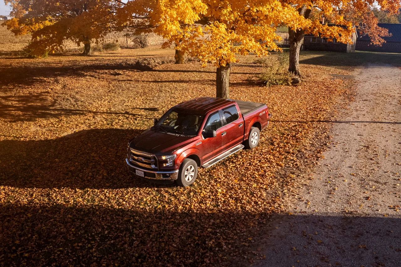 A red pickup sits beneath golden autumn trees, casting a long shadow.