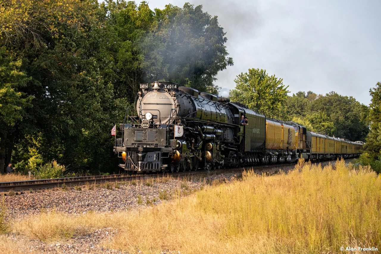 A steam locomotive emerges beside golden grass with smoke rising into the sky.